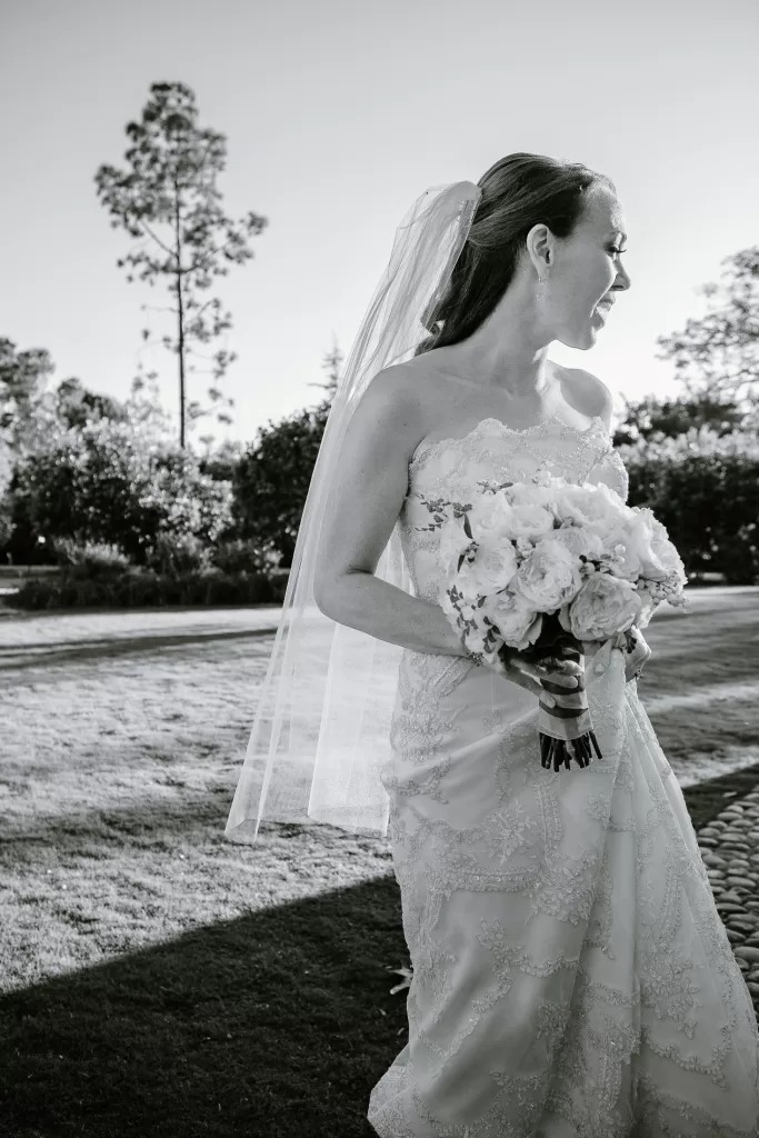 A woman in a strapless bridal gown with lace details holds a bouquet of flowers and stands outdoors on a sunny day. She has a veil and smiles while looking to the side. The background features grass, trees, and bushes. The image is in black and white.