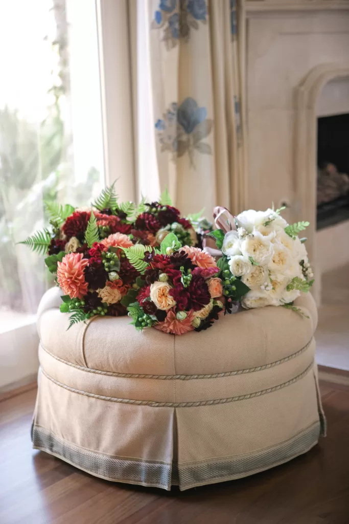 A beige upholstered ottoman is adorned with an assortment of floral bouquets featuring rich autumnal colors, including deep reds, peach, and white, with greenery interspersed. Background includes floral curtains and part of a fireplace.