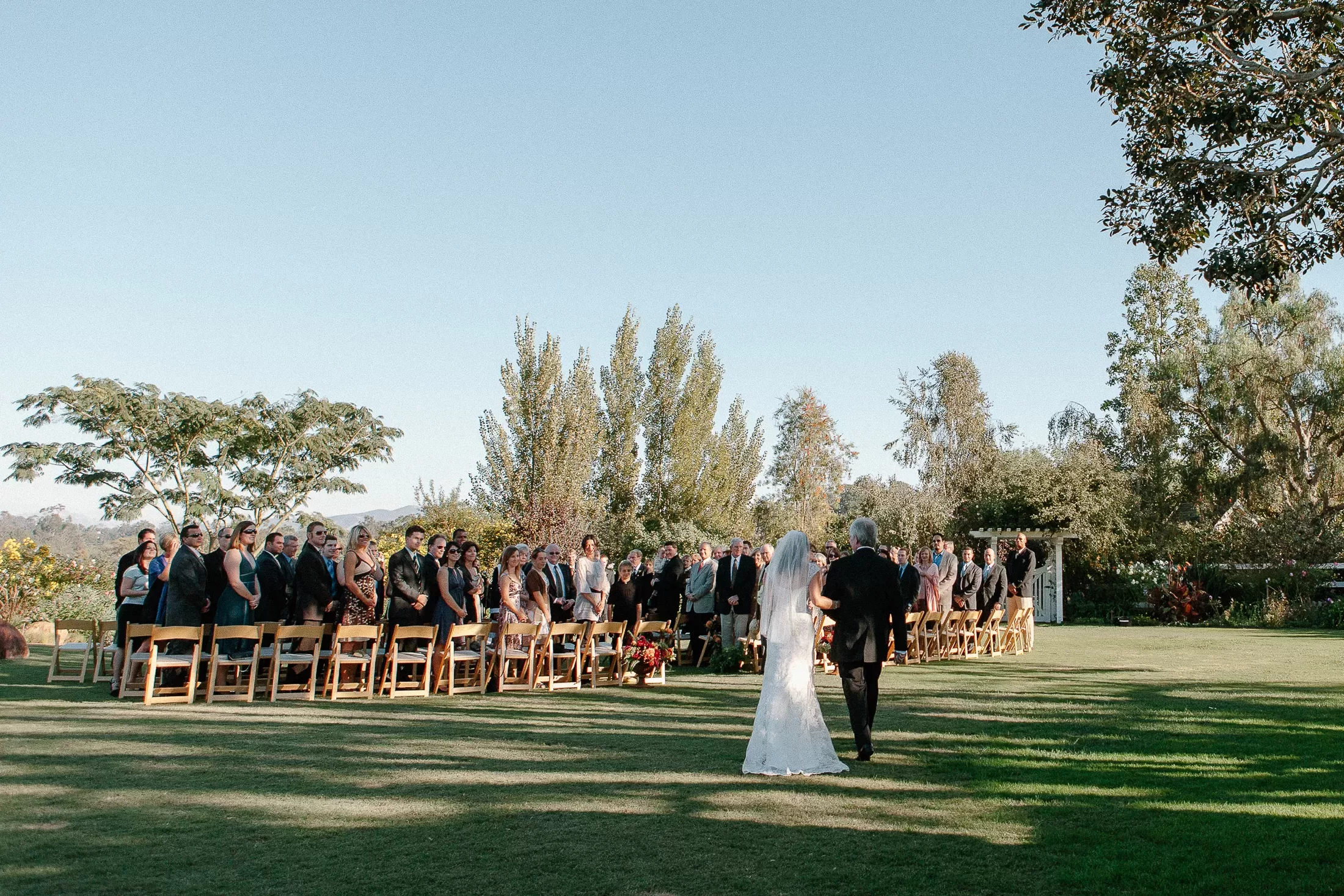 A bride, wearing a white wedding dress and veil, walks down an outdoor grassy aisle accompanied by an older man in a suit. Guests seated on wooden chairs on either side of the aisle turn to watch them. Trees and a clear blue sky form the background.