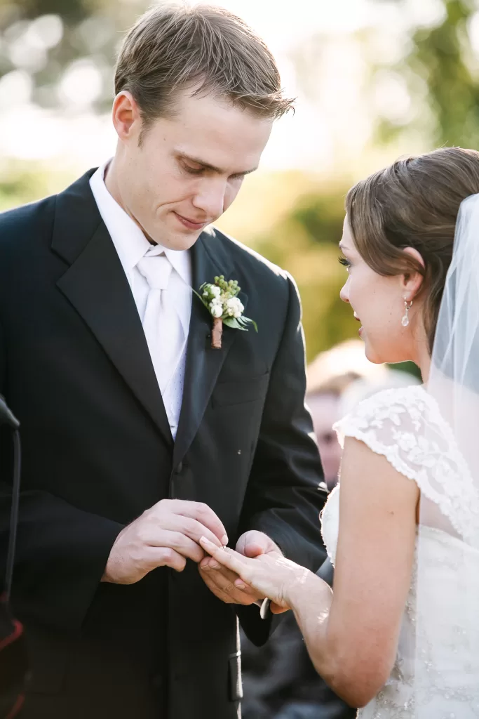 A groom in a black suit and white tie places a ring on the bride's finger during an outdoor wedding ceremony. The bride, wearing a white lace dress and veil, looks at the groom. They are surrounded by greenery and bathed in soft sunlight.