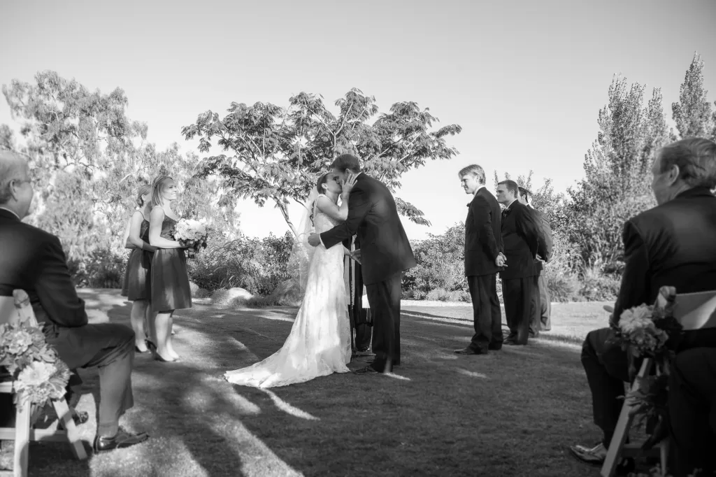 A bride and groom share a kiss during an outdoor wedding ceremony. The bride is in a long white dress, and the groom is in a dark suit. Bridesmaids and groomsmen stand nearby, while guests sit on either side watching the couple. Trees and greenery surround them.