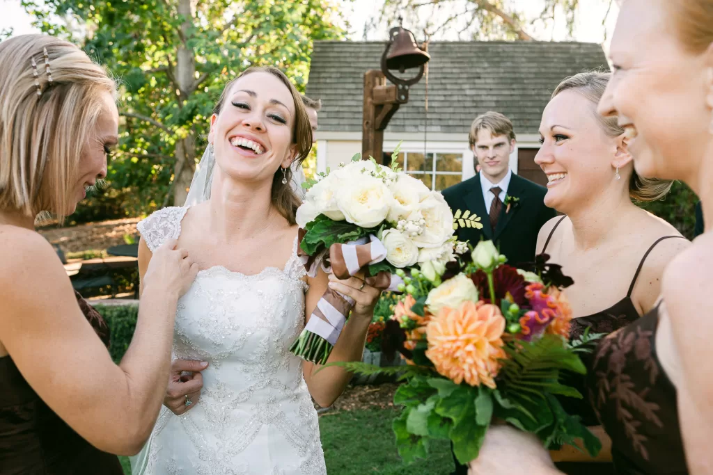 A smiling bride in a white dress stands outdoors holding a bouquet of white flowers. She is surrounded by three bridesmaids in brown dresses who are also holding bouquets and smiling. A man in a suit stands in the background. Trees and a building are visible behind them.