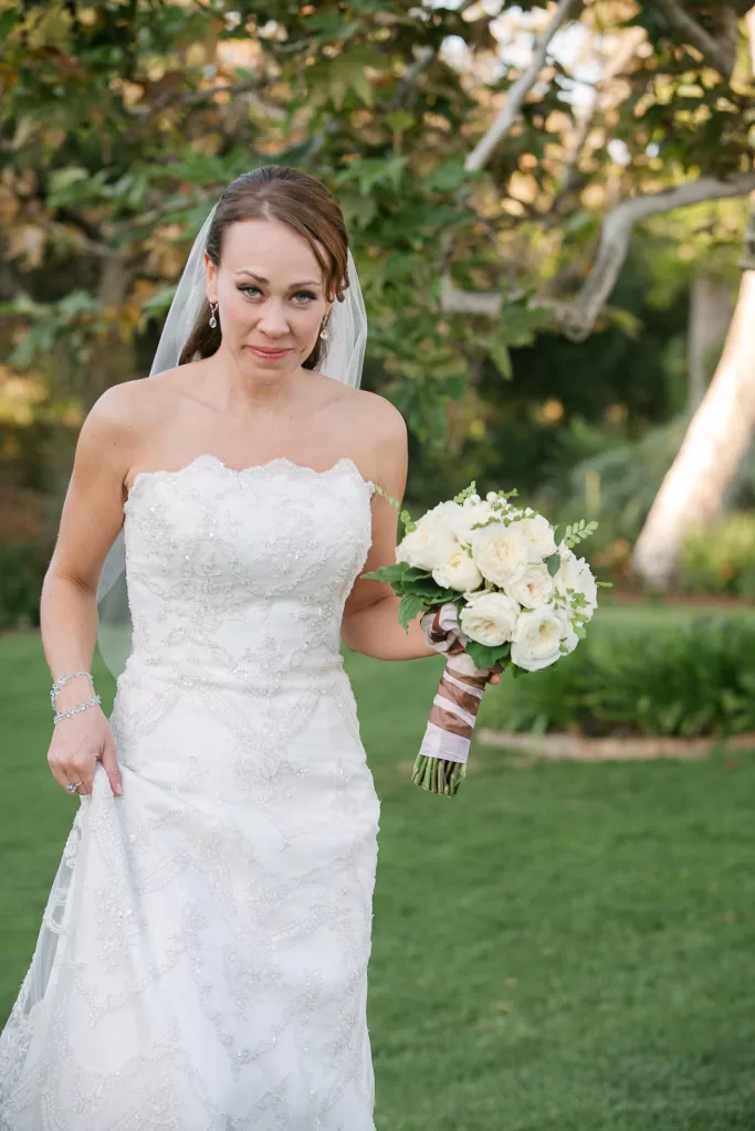 A bride in a strapless white lace wedding dress holds a bouquet of white flowers wrapped in ribbon. She stands on green grass with trees in the background, wearing a veil and a bracelet, and looks at the camera with a slight smile.