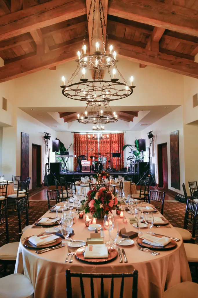 An elegantly decorated Rancho Santa Fe Country Club set for a formal event. A round table with place settings, floral centerpieces, and candles is in the foreground. In the background, a stage with musical instruments and a backdrop of long curtains is visible under two large chandeliers.