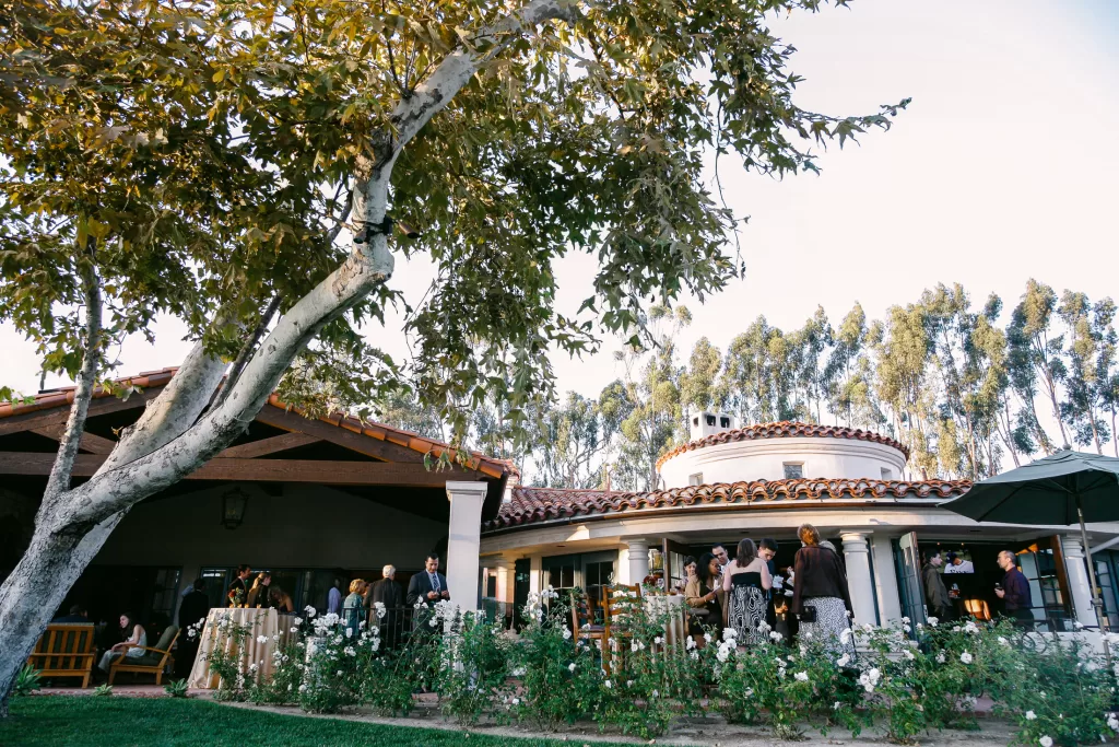 A vibrant outdoor wedding reception at the picturesque Rancho Santa Fe Country Club shows a white circular building and a large tree. Guests mingle under a wooden pergola and amongst lush green gardens with blooming flowers. The sky is clear, adding to the scenic atmosphere.