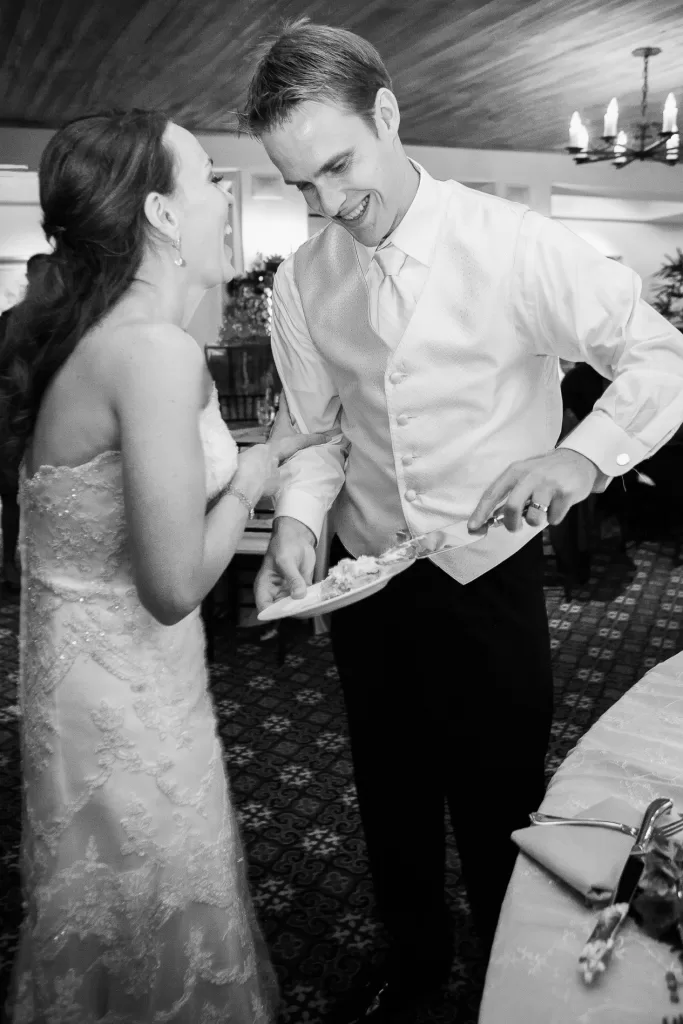 A bride and groom are standing closely, both smiling and laughing. The groom is holding a plate and a piece of cake, wearing a white vest and tie. The bride is in a strapless, embellished gown. The setting appears to be indoors, in a warmly lit room with a chandelier.