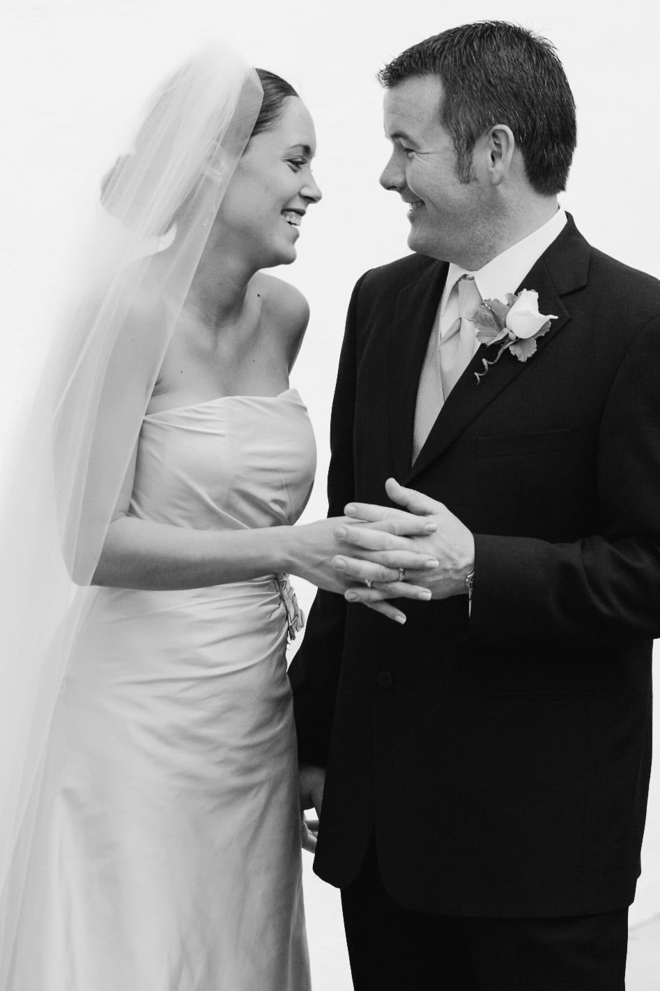 A bride and groom stand together, smiling at each other. The bride is wearing a strapless gown and veil, while the groom is in a suit with a boutonniere. They are holding hands and appear happy and joyful.