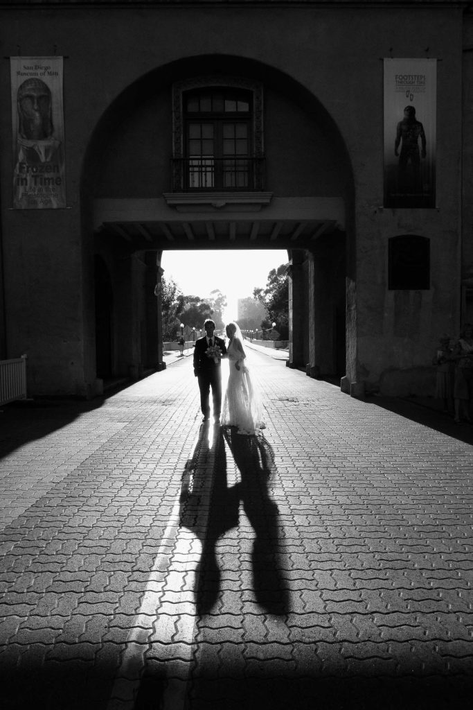 A bride and groom holding hands walk through a sunlit arched passageway, casting long shadows on the brick path. The scene is in black and white, adding a timeless feel. Two banners hang on either side of the arch.