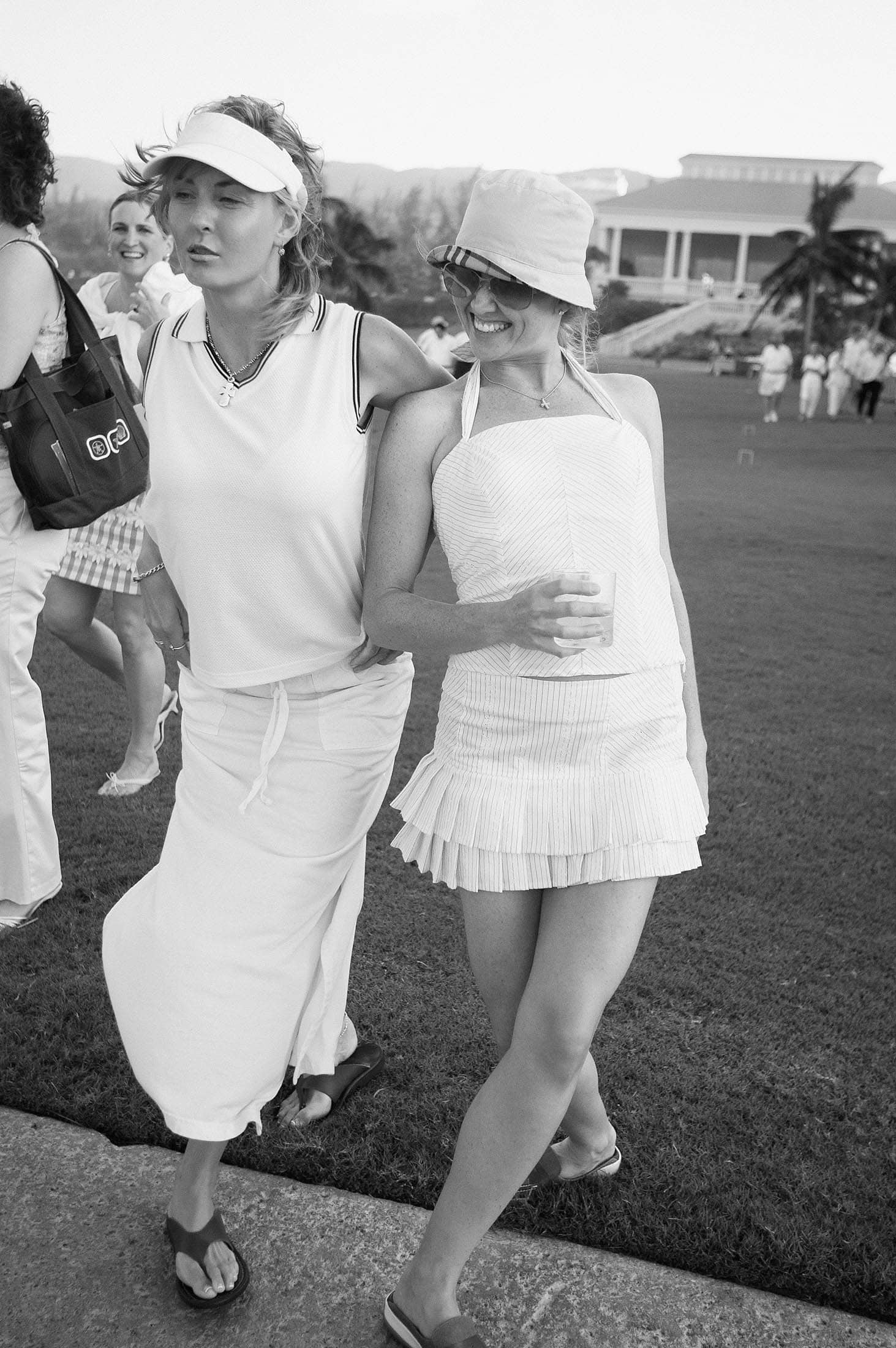 Two women in summer outfits walk arm in arm on a golf course. One wears a visor and long skirt, while the other wears a sunhat and short skirt. People and a large building are visible in the background. They're smiling and seem relaxed.