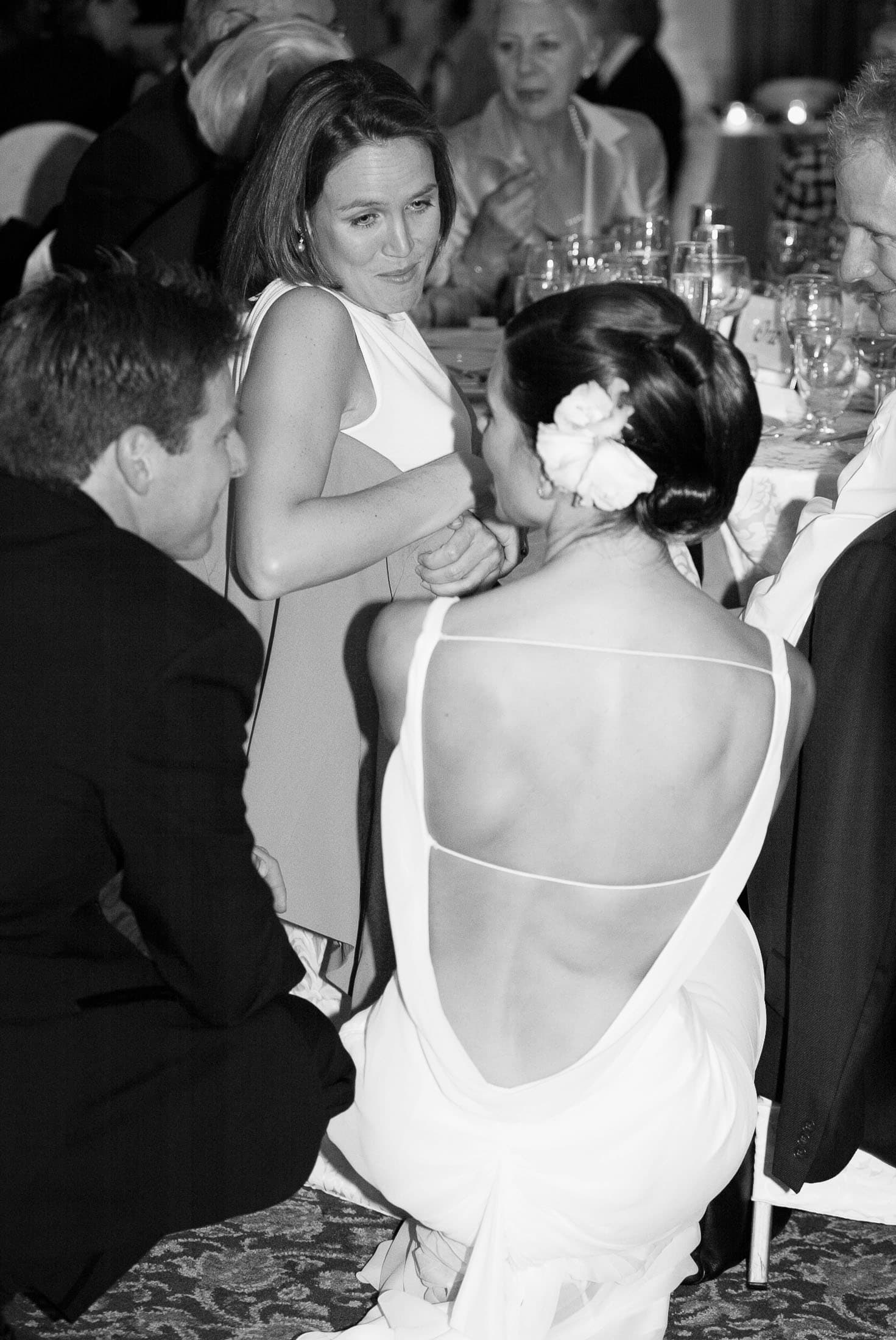 A black-and-white photo of people at a formal event. A woman in a backless dress is seated, facing a woman in a sleeveless dress. Two men in suits are nearby. They're engaged in conversation at a table with glasses and tableware.