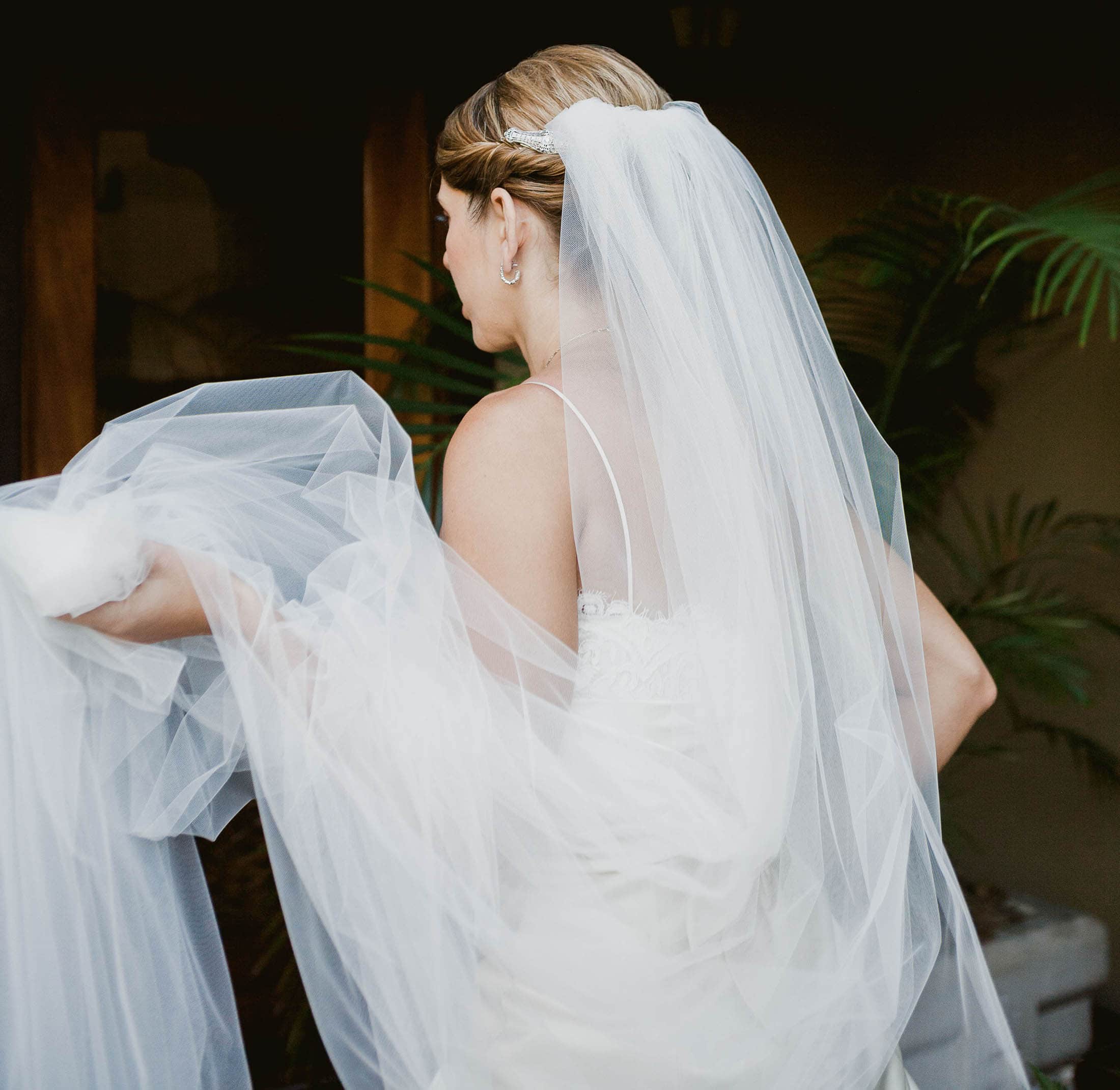 A bride in a white dress and veil is seen from the back, holding up her gown. She stands near a plant, with soft lighting highlighting her elegant attire and hairstyle.