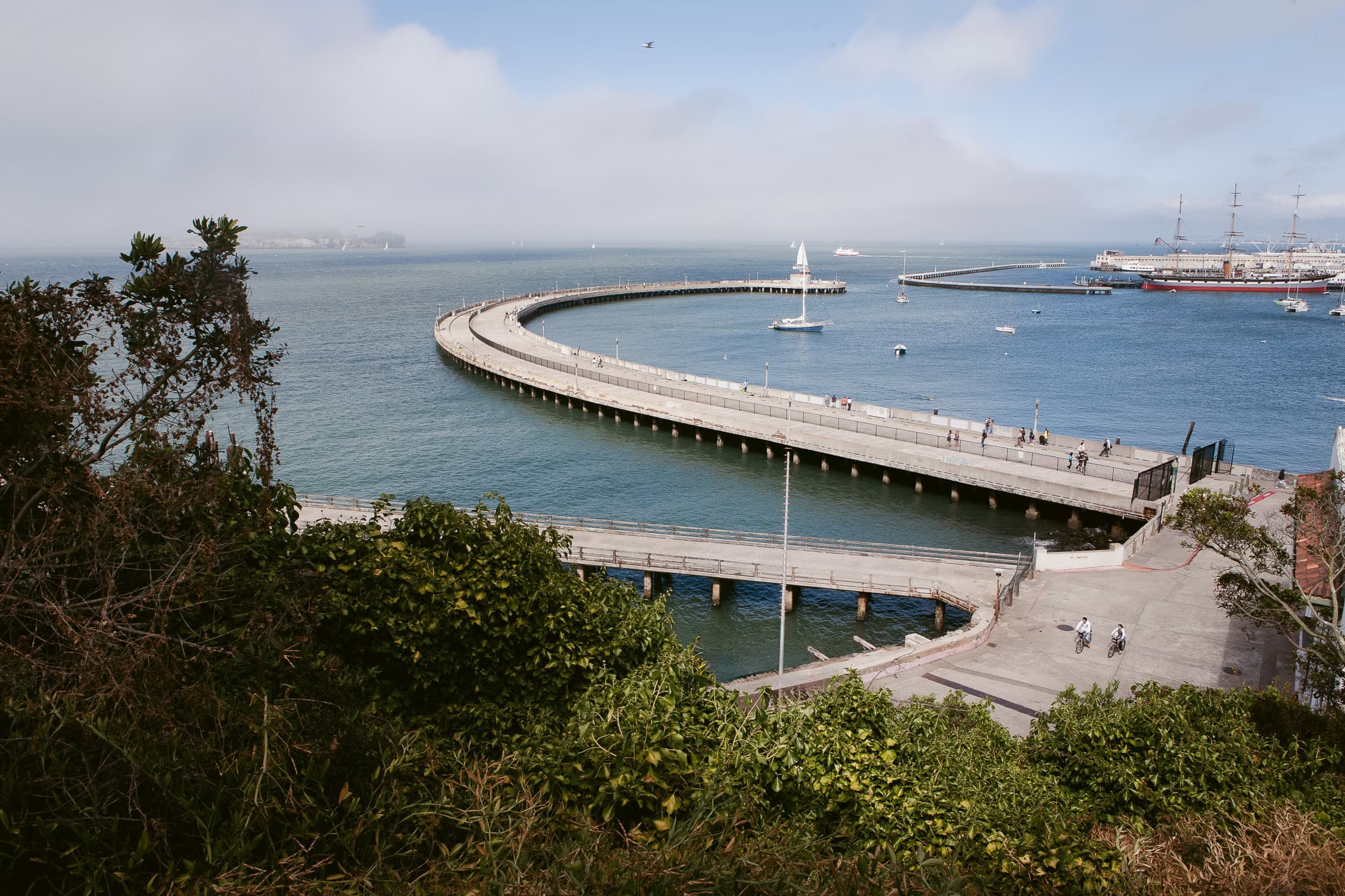 A scenic view of a curving pier extending into the bay offers a perfect setting for San Diego engagement photos, with people strolling along it. The blue waters are calm under a partly cloudy sky, boats and a large ship dotting the distance, while lush greenery frames the foreground.