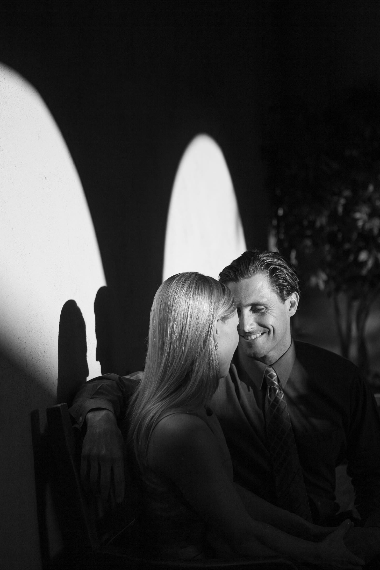 A black and white photo captures a couple sitting closely on a bench during their San Diego engagement, facing each other with smiles. Light and shadows create an intricate pattern on the wall behind them. The man is in a suit and tie, while the woman, with her long hair, exudes elegance.
