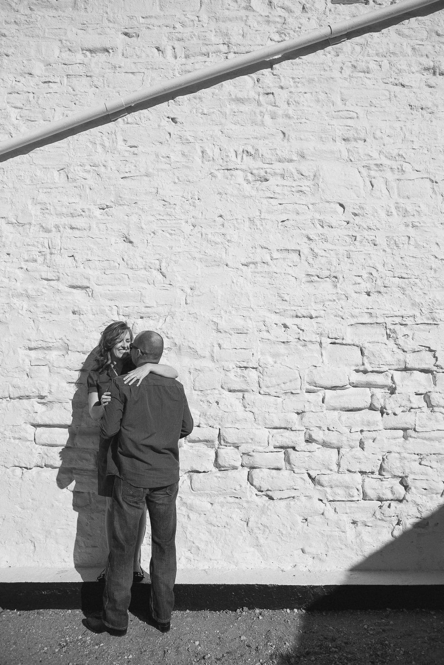 A couple stands close together against a sunlit brick wall, capturing the essence of San Diego engagement photos. The woman leans back, smiling up at the man. The scene is in black and white, casting soft shadows on the ground.