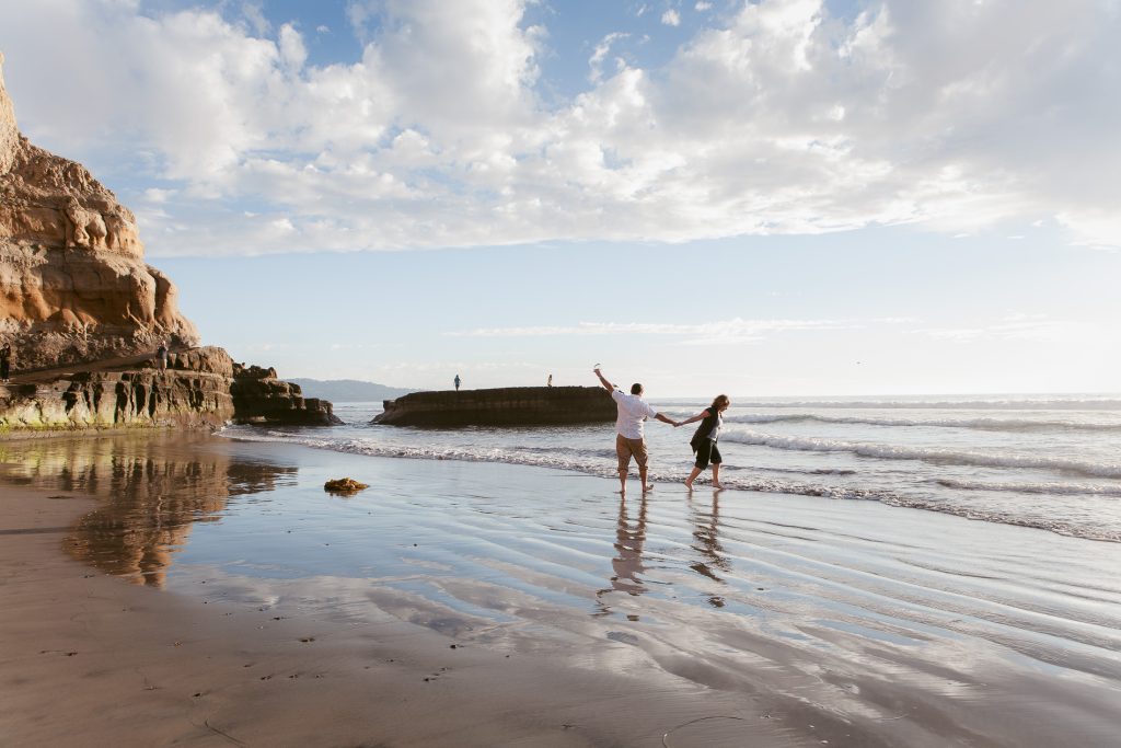 A couple holds hands while walking along a scenic beach at sunset, perfect for San Diego engagement photos. They appear joyful as they enjoy the water and sand, with cliffs and a cloudy sky enhancing the serene atmosphere.
