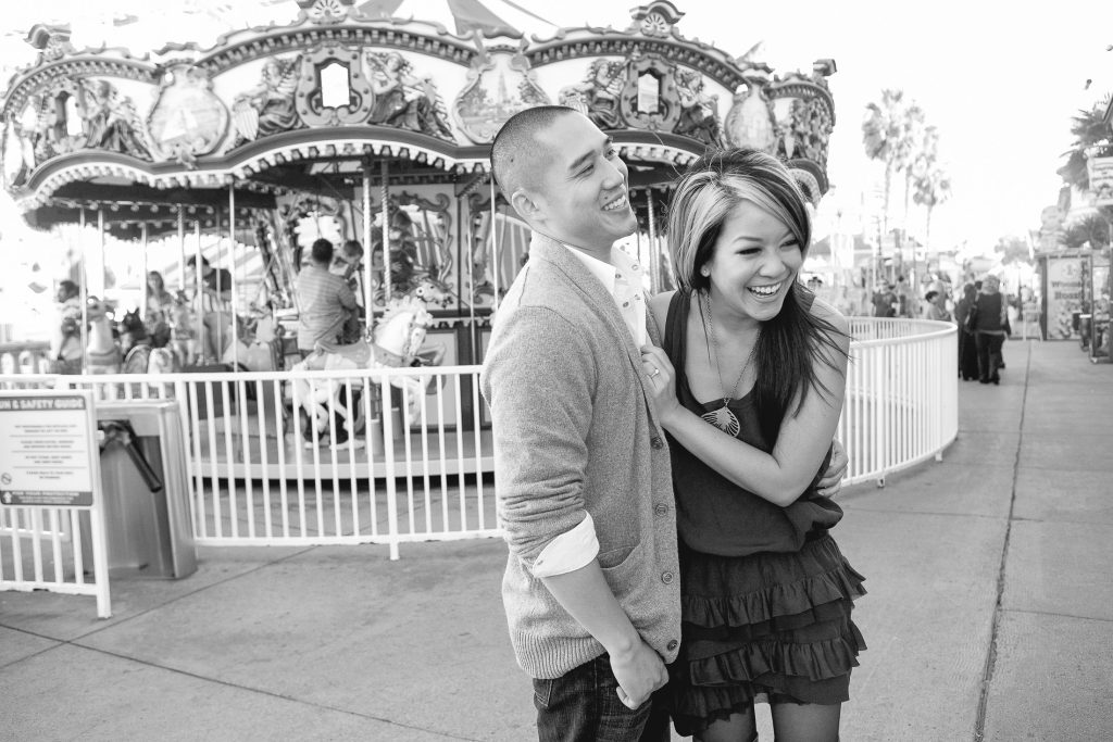 A young couple joyfully embraces at a vibrant San Diego fairground, perfect for engagement photos. The woman is in a dress and the man wears a cozy sweater, with a colorful carousel behind them and lively crowds enjoying the attractions.