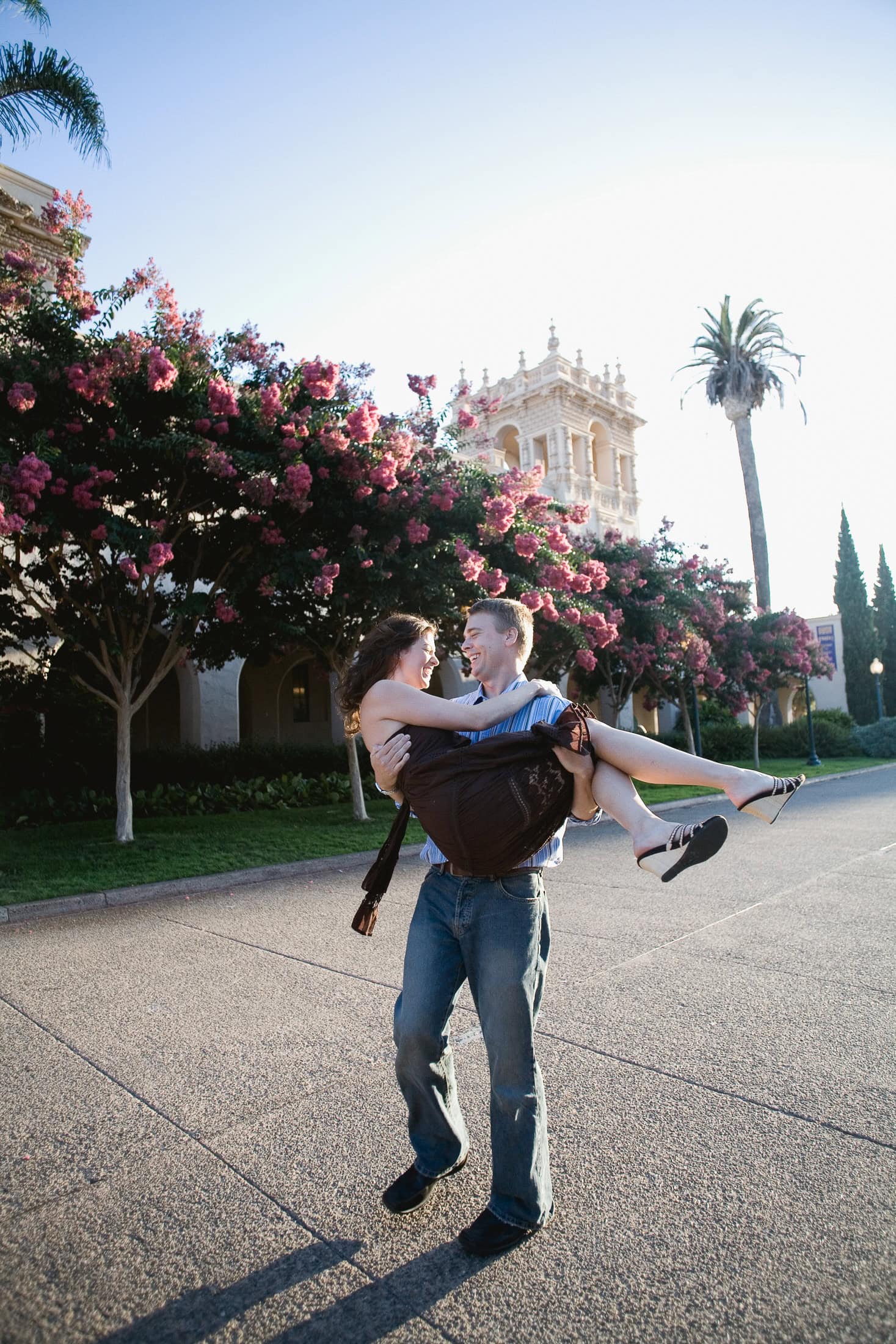 A man joyfully lifts a woman in his arms on a sunny day, capturing the essence of San Diego engagement photos. They're surrounded by blooming trees with a historic building in the background, appearing happy and carefree.