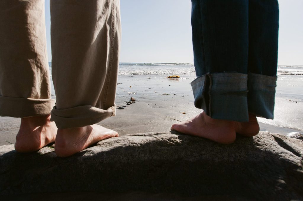Two people stand barefoot on a rock at the beach, capturing the essence of San Diego engagement photos. The ocean and sandy shore paint a serene backdrop, while their slightly rolled-up pant legs add to the relaxed, casual atmosphere.