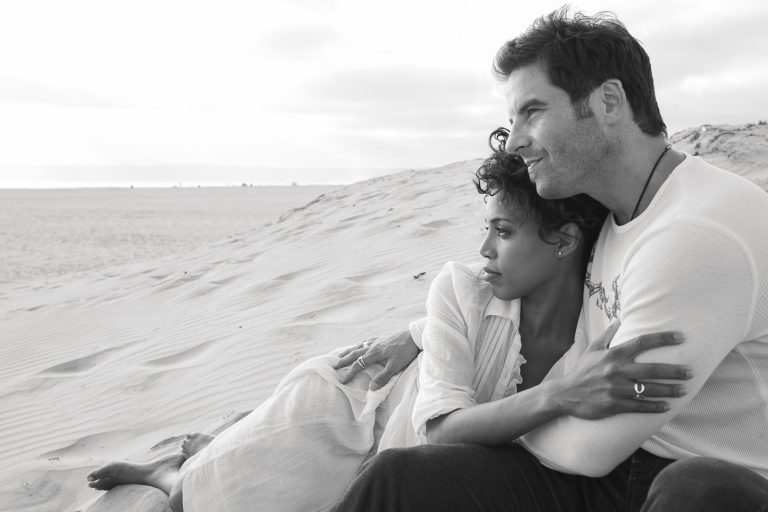 A couple sits together on a sandy beach, embracing while gazing at the horizon. The woman leans on the man's chest, their San Diego engagement photos capturing this timeless, serene scene in black and white.