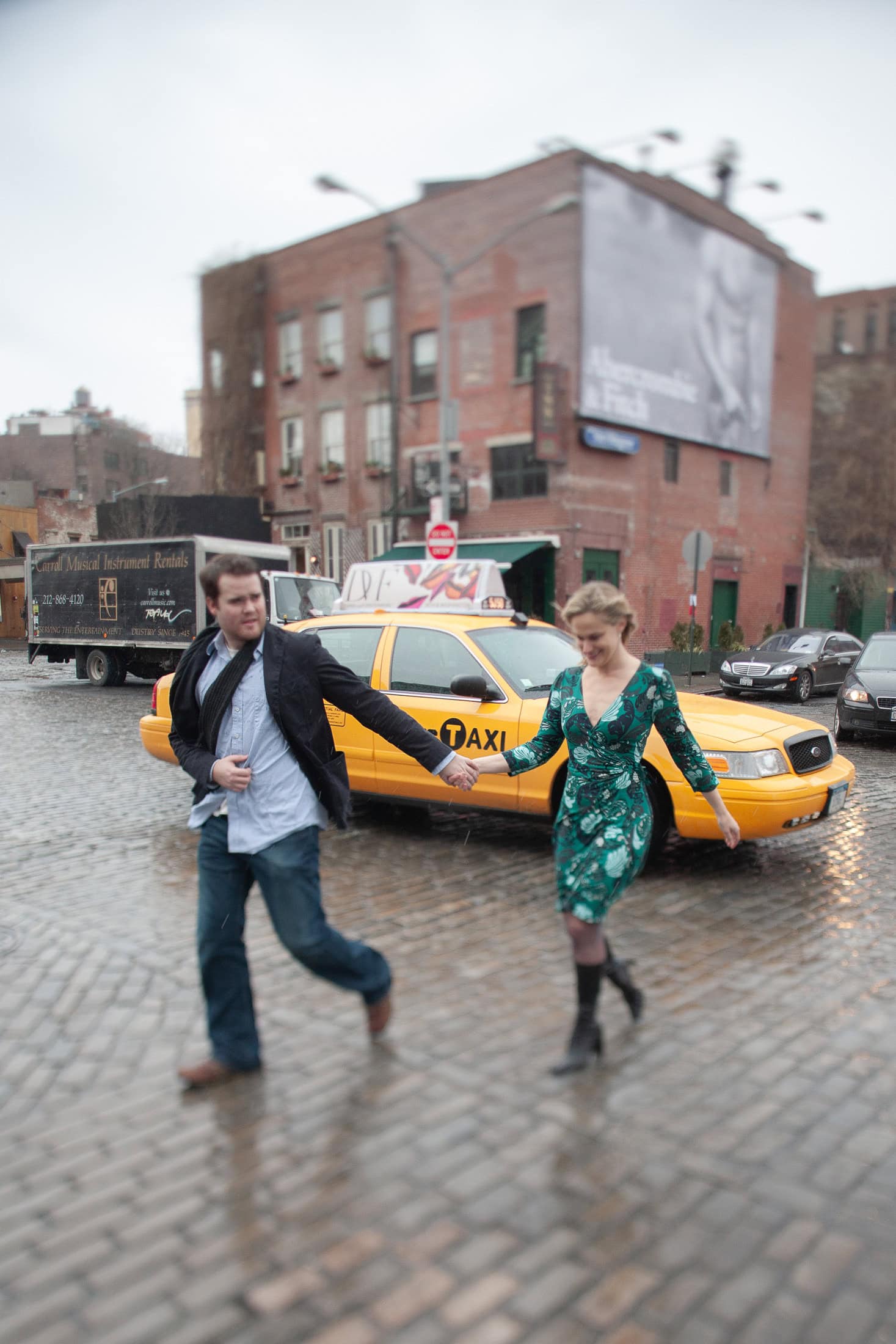 A couple holds hands while crossing a wet cobblestone street, capturing the essence of San Diego engagement photos. A yellow taxi and red brick building form the backdrop as she elegantly wears a green dress, both appearing in motion.