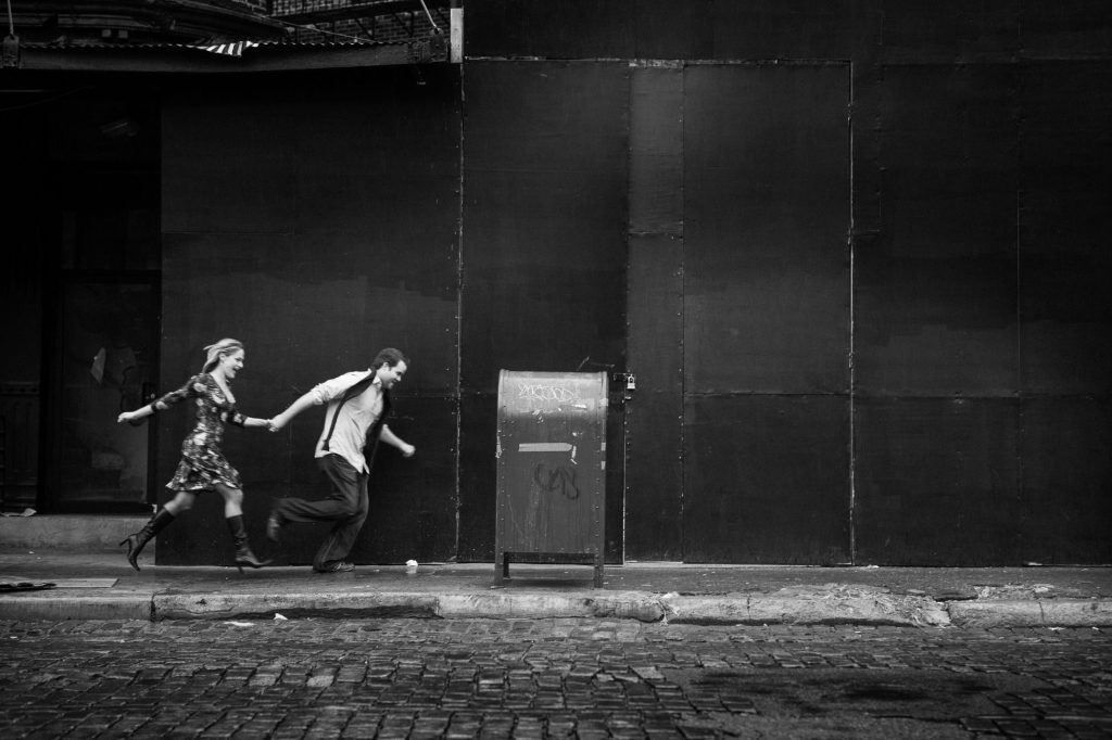 A black and white photo captures a couple holding hands, running on a cobblestone street in San Diego past a mailbox. The urban backdrop with its large dark wall adds to the engagement photo's sense of urgency and spontaneity.