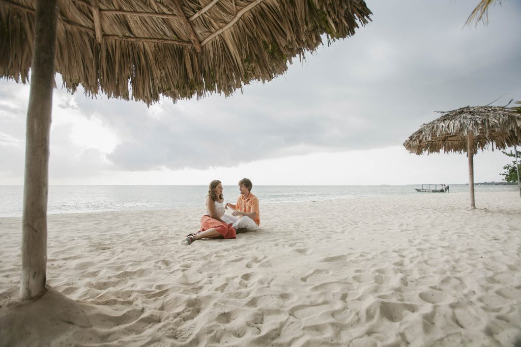 Negril, Jamaica: A couple sits together under a thatched umbrella on a sandy beach, facing each other and smiling, with the ocean and a cloudy sky in the background. Another thatched umbrella and a boat are visible in the distance.