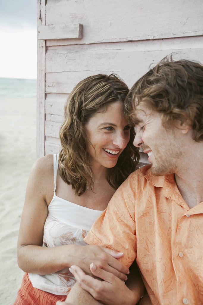 A smiling couple sits close together by a light pink wooden beach hut, looking at each other lovingly. The beach and ocean are visible in the background.