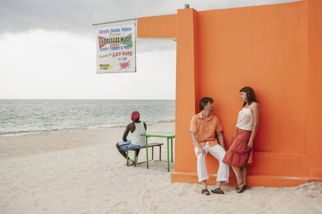 Negril, Jamaica: A man and woman in casual summer clothes lean against a bright orange wall on a sandy beach, smiling at each other. Nearby, another person in a red headscarf sits at a table facing the ocean. A sign advertises live reggae music.