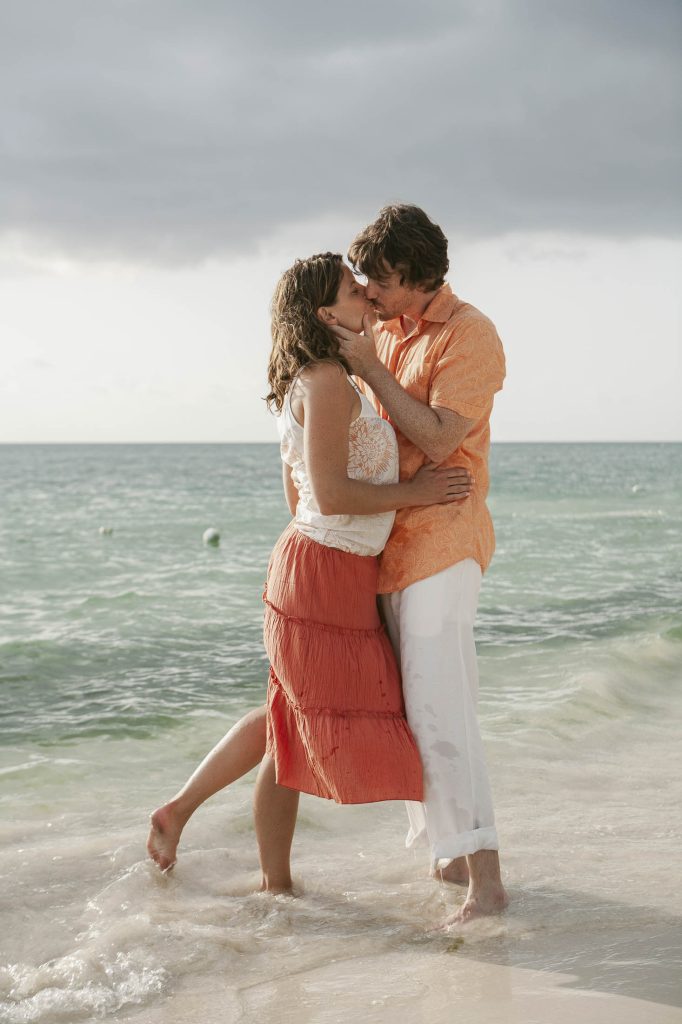 Negril, Jamaica: A couple stands on the beach at the edge of the water, embracing and kissing. They are both dressed in light, summery clothes, and the ocean waves and cloudy sky form the background.