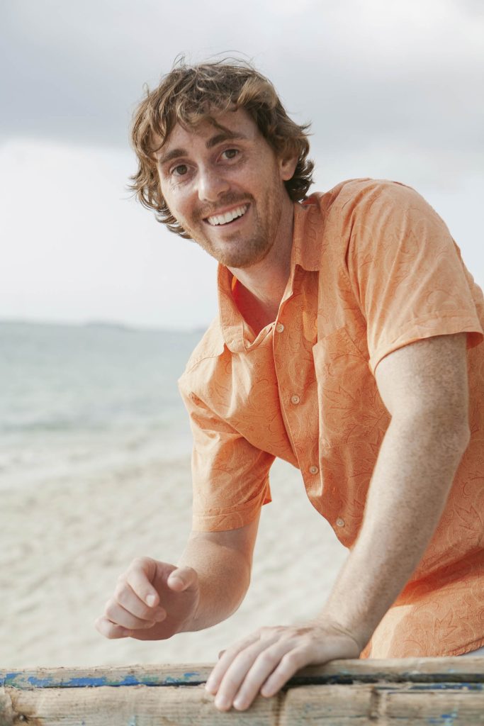 A smiling man with wavy brown hair wearing an orange short-sleeve shirt stands on a sandy beach with the ocean and a cloudy sky in the background.