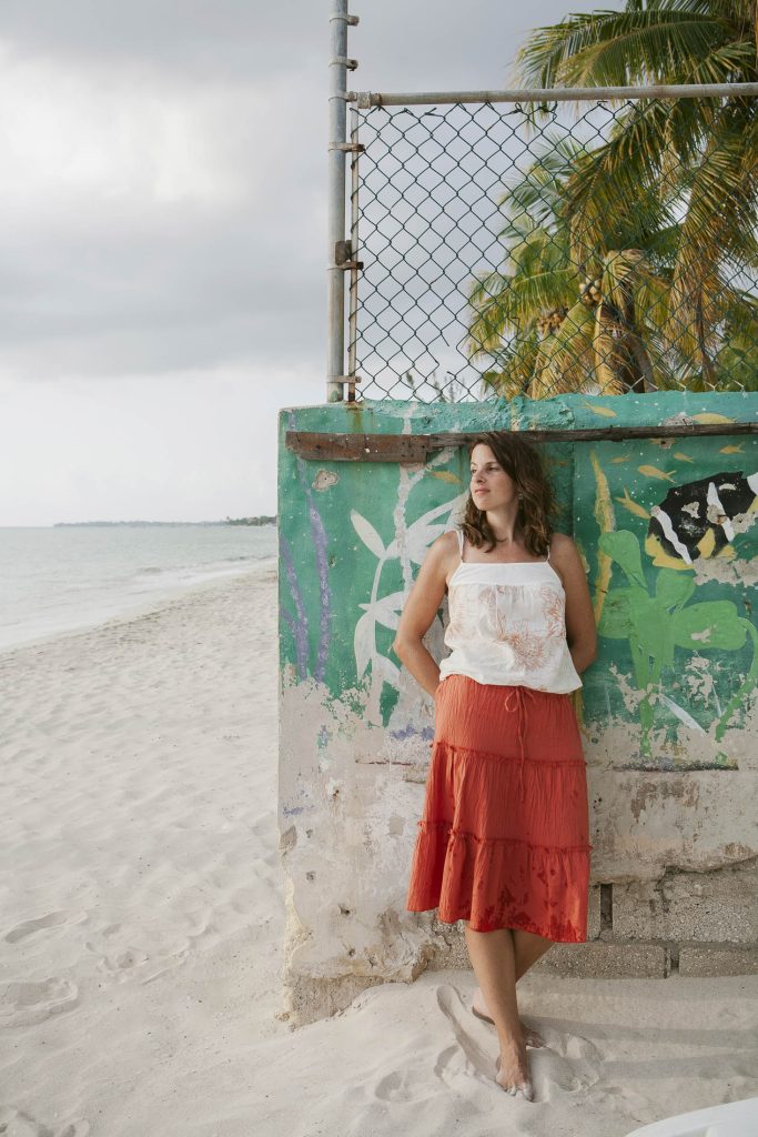 A woman in a white top and orange skirt stands barefoot on a sandy beach, leaning against a colorful, weathered wall with palm trees and ocean in the background.