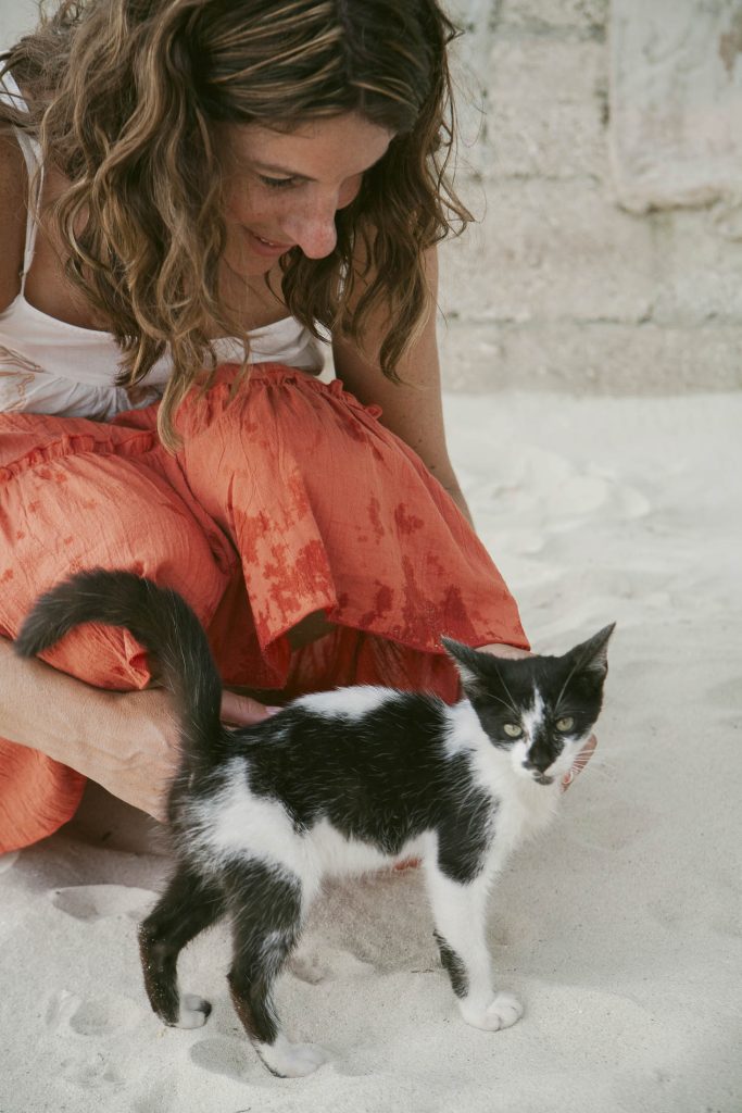 A woman in an orange skirt smiles down at a black and white cat standing on white sand, gently petting its back.