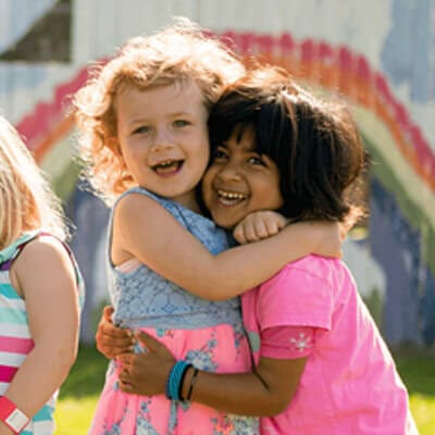 Two young children hugging while smiling. There is a rainbow painted on the wall behind them.