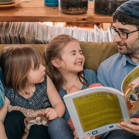 A man reading a book to two children. The child in the middle is smiling with their eyes closed.