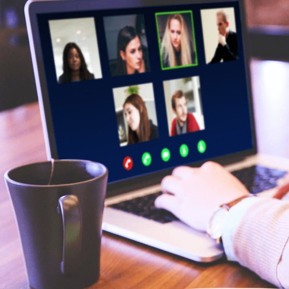 A close up of a person on a video call on their laptop. There is a brown mug in focus to the left of the computer.