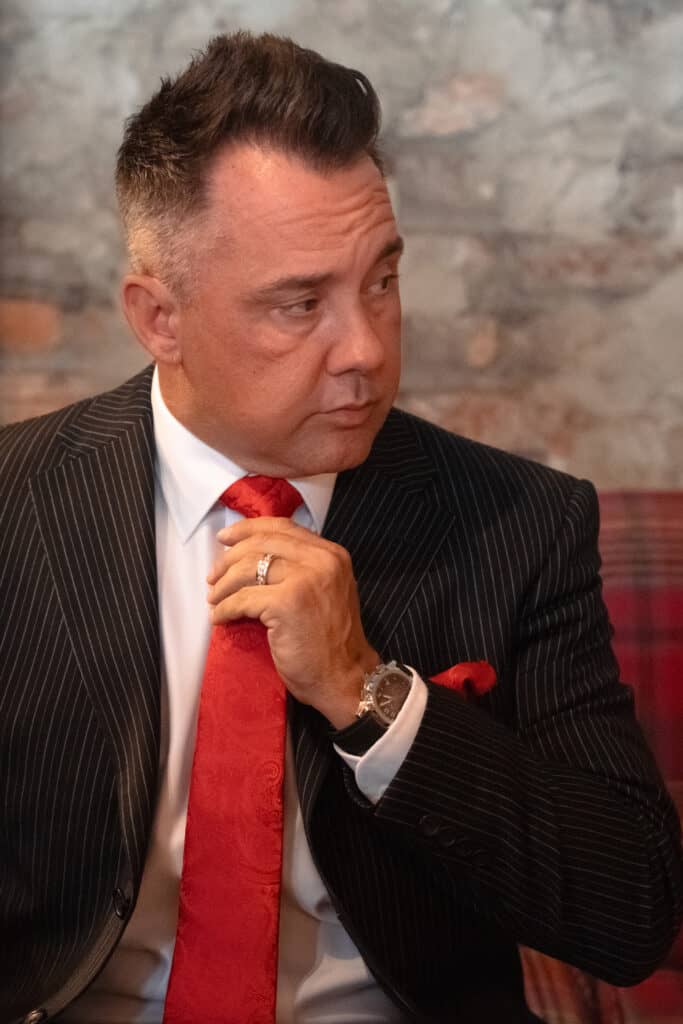 Professional businessman in formal suit and red tie, sitting with hand on chin.