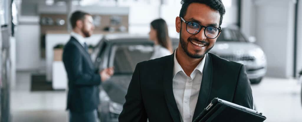 Professional businesswoman smiling at car dealership, holding clipboard in showroom, happy customer service, automotive sales, branded dealership environment.