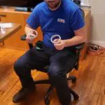A person in a blue shirt sits on an office chair, holding two rolls of adhesive tape. The room has wooden flooring and a desk in the background.