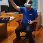 Person seated in an office wearing a virtual reality headset and holding a VR controller, gesturing upwards with one hand.