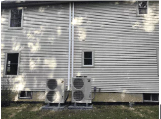 Two outdoor air conditioning units are installed on the ground outside a light-colored siding building, with two small windows visible above.
