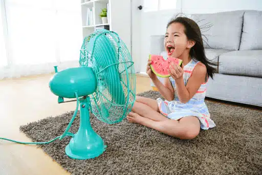 A girl sits on a rug, enjoying watermelon, while a turquoise fan gently blows her hair in the living room. It's as refreshing as reading a glowing customer review on a hot summer day.