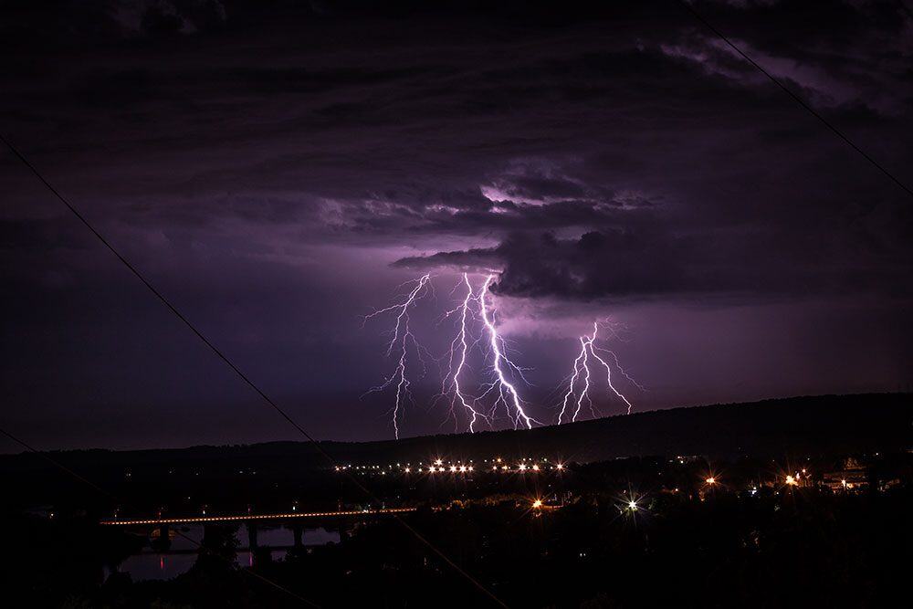 Lightning illuminates a stormy sky over a distant cityscape at night, with visible power lines and a glowing horizon.