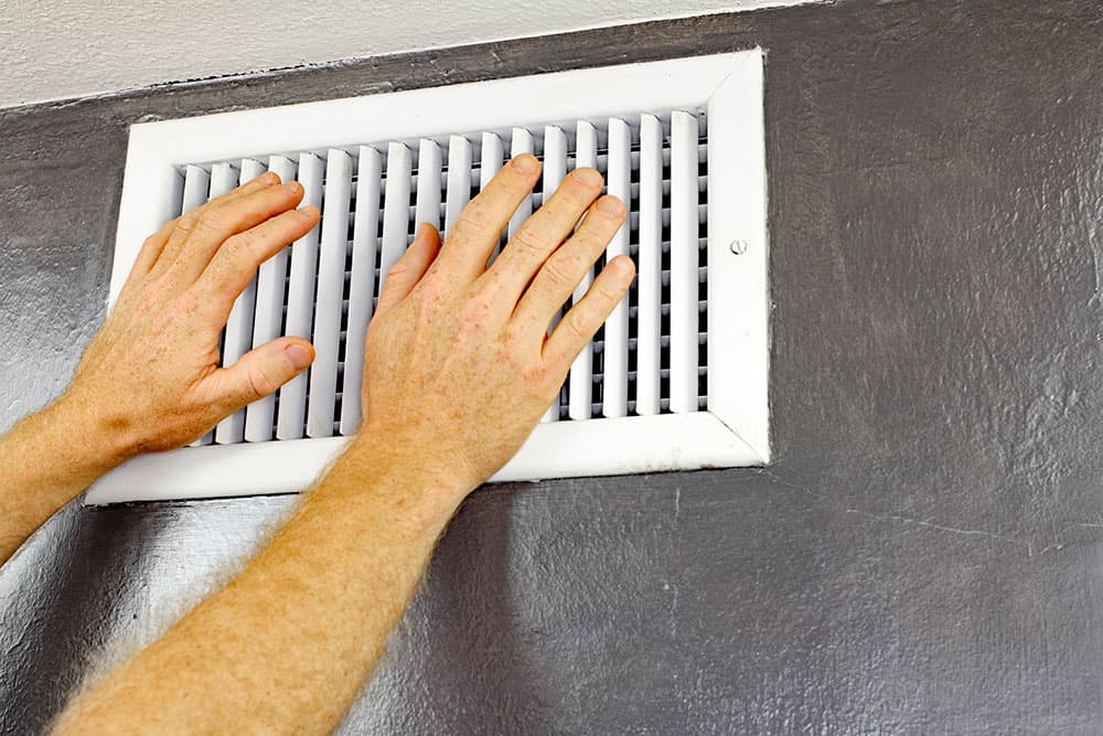 Two hands are held up to a white wall vent, which is mounted on a dark gray wall—an example of affordable basement heating in action.