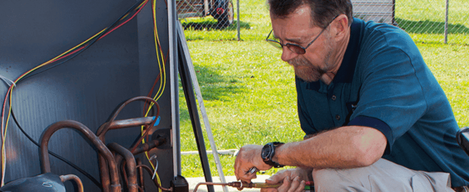 Person in a blue shirt examines and adjusts equipment with wires and copper pipes outdoors on a sunny day.