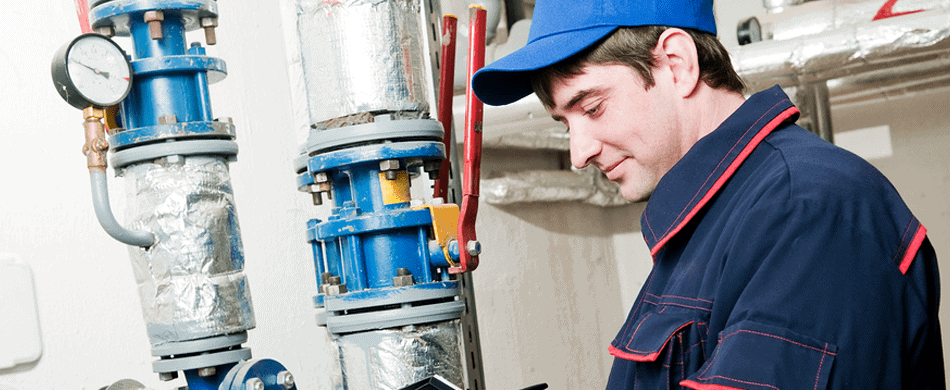 A worker in a blue cap and uniform checks pressure gauges and piping in an industrial setting.
