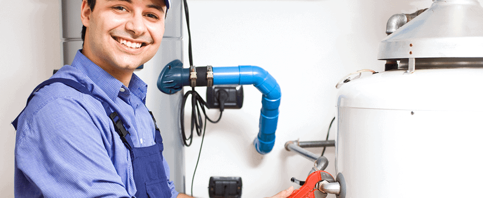 A smiling technician in a blue uniform adjusts a valve on a water heater with a wrench.