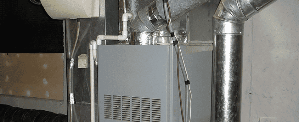 A gray furnace with metal ducts and white pipes is installed in a basement. The wall is partially unfinished, showing exposed drywall.