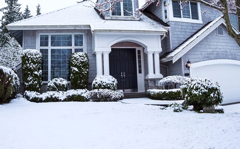 A gray house with white trim and double doors stands surrounded by snow-covered bushes, lawn, and trees in winter—reminding us that heating in cold weather is essential for comfort and safety.