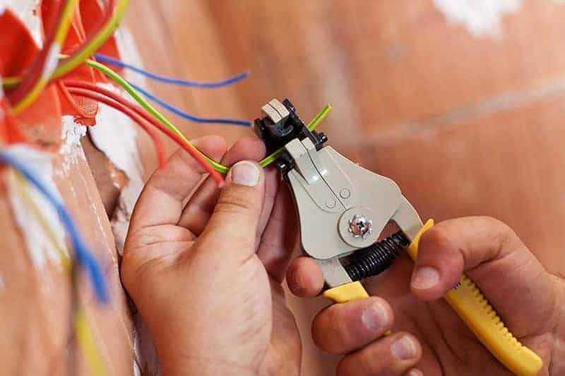 Close-up of hands using wire stripper tool to remove insulation from electrical wires protruding from a wall socket.