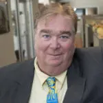 Man wearing a suit with a yellow shirt and blue patterned tie, smiling indoors.